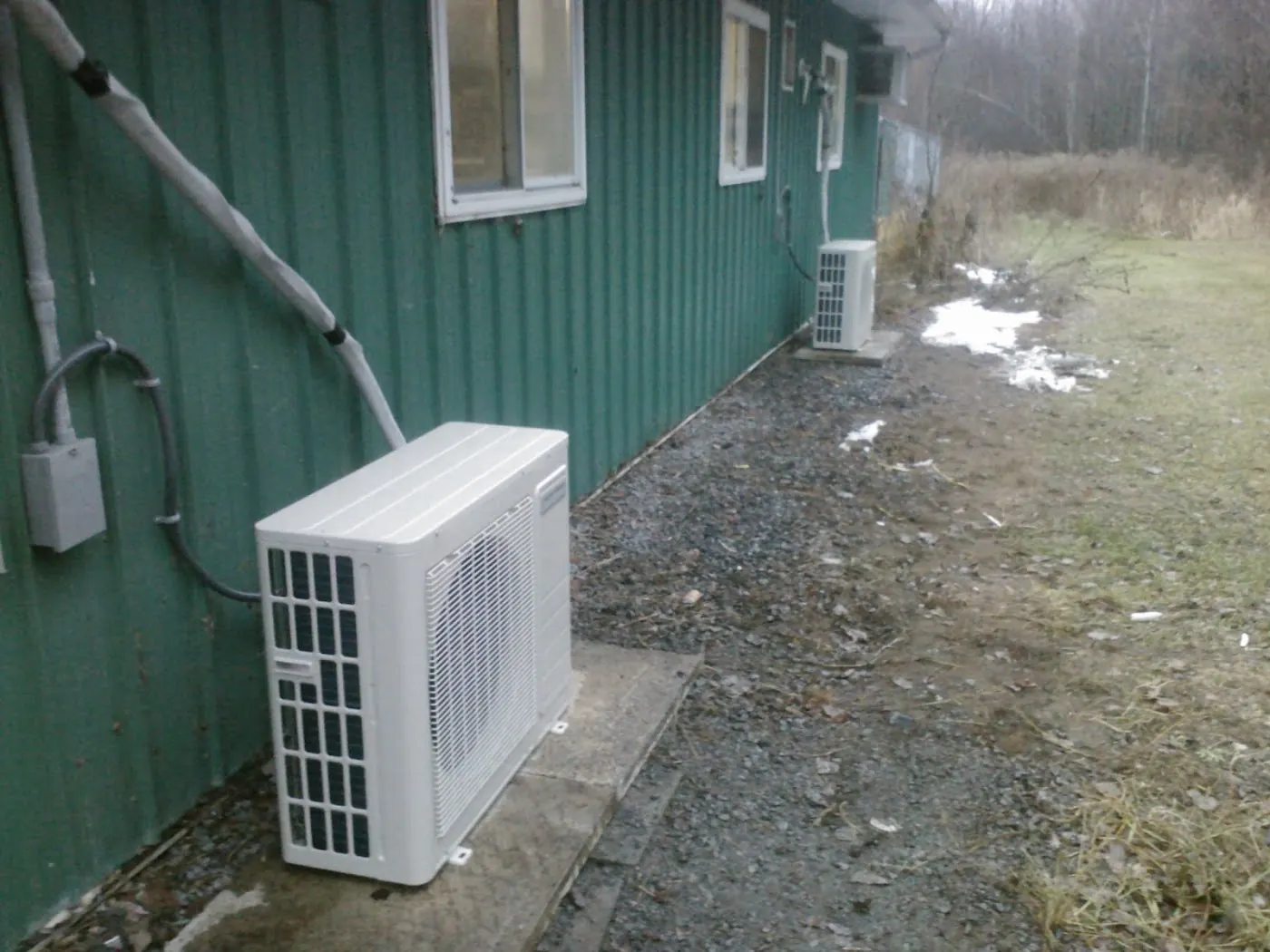 Two outdoor condenser units along a building with green metal siding